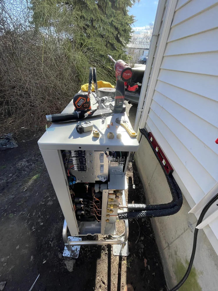 An outdoor unit of an HVAC system is being installed next to a white house. Various tools, including a drill, tape measure, and wrenches, are neatly laid out on top of the unit.